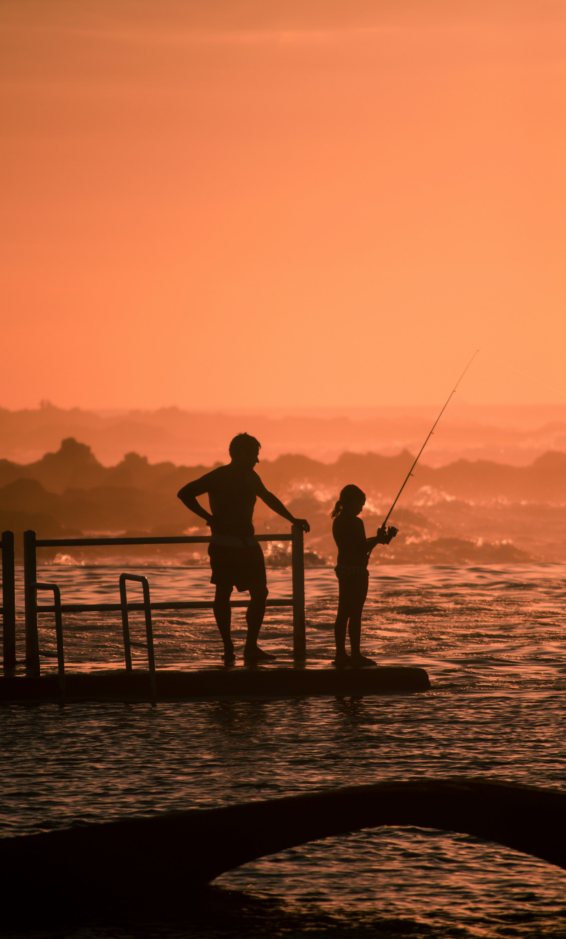 A father and daughter fishing side by side, silhouetted against the sunset